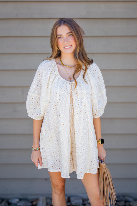 Ivory floral eyelet romper with puff sleeves, front tie detail, and flowy babydoll fit styled with gold jewelry and a neutral fringe handbag for a summer boutique outfit.