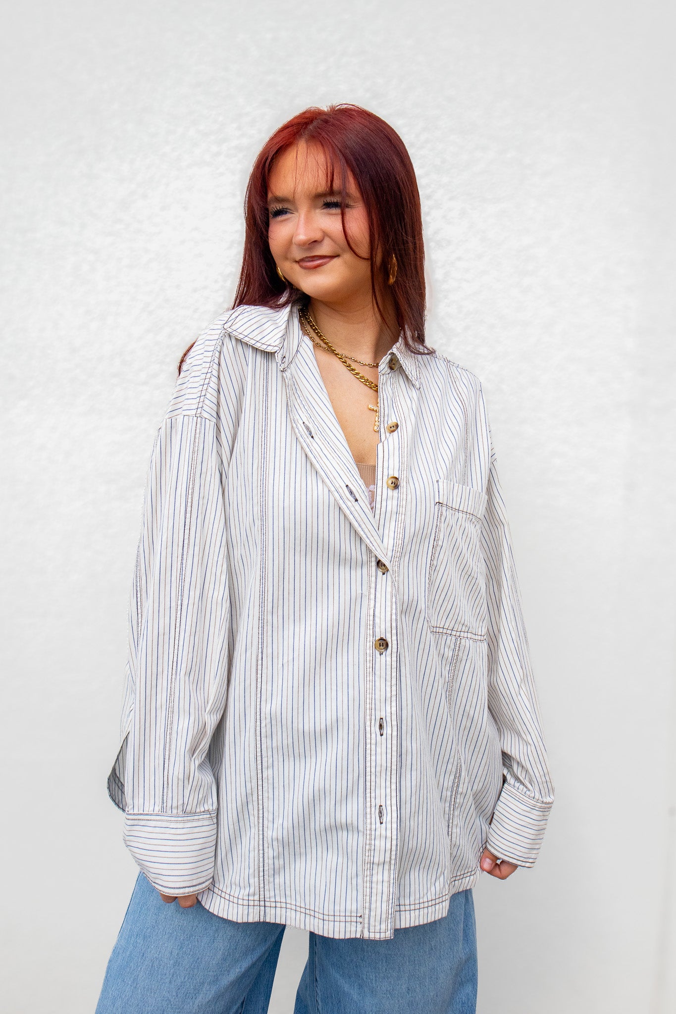 A woman with red hair wearing an oversized white button-up shirt with thin vertical blue stripes, paired with light blue jeans, standing against a textured white wall.