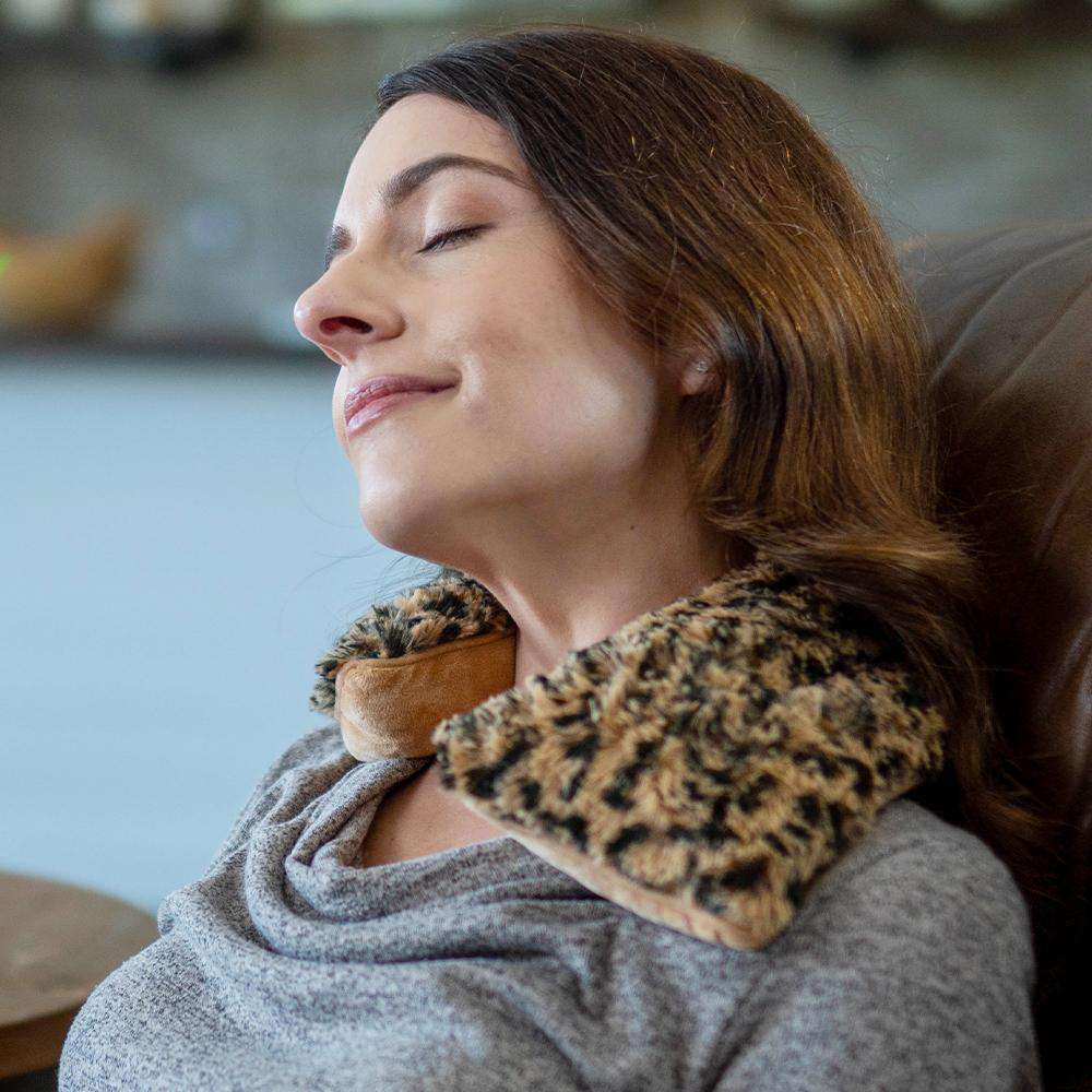 Woman relaxing in a chair with a plush leopard print Warmies® neck wrap draped around her shoulders, providing soothing heat therapy and cozy comfort for neck and shoulder relief.