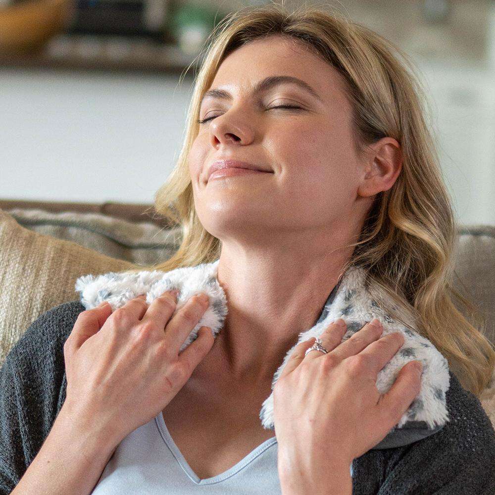 Woman relaxing with a plush Warmies® neck wrap draped around her shoulders, providing soothing heat therapy and cozy comfort for neck and shoulder relief.