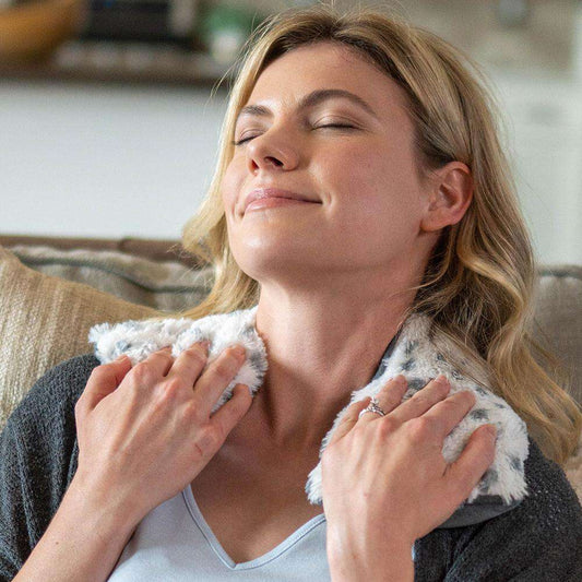 Woman relaxing with a plush Warmies® neck wrap draped around her shoulders, providing soothing heat therapy and cozy comfort for neck and shoulder relief.