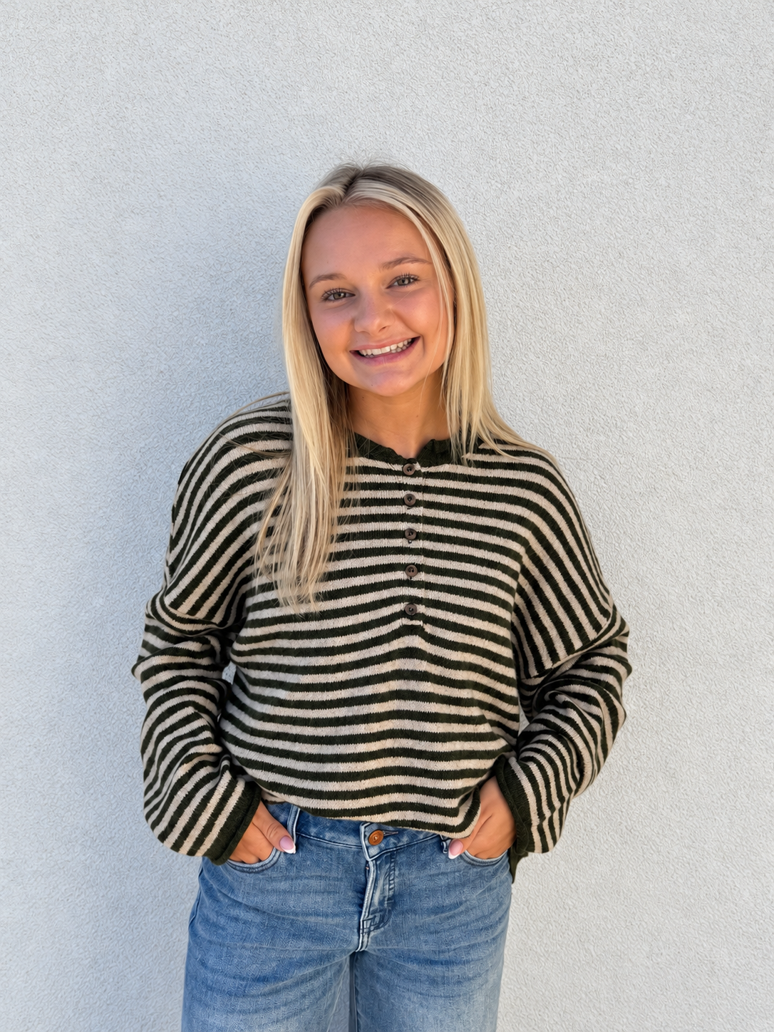 Model wearing the Weekend Stripe Henley Sweater in Olive Green/Cream, featuring classic horizontal stripes, a relaxed fit, soft knit fabric, button-front neckline, and long drop-shoulder sleeves, styled with light-wash denim jeans.