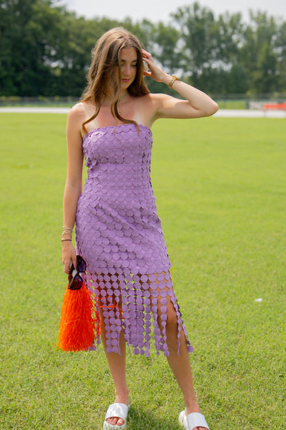 A woman stands on a grassy field wearing a lavender strapless crochet dress with a fringe hem, holding a orange pom pom.