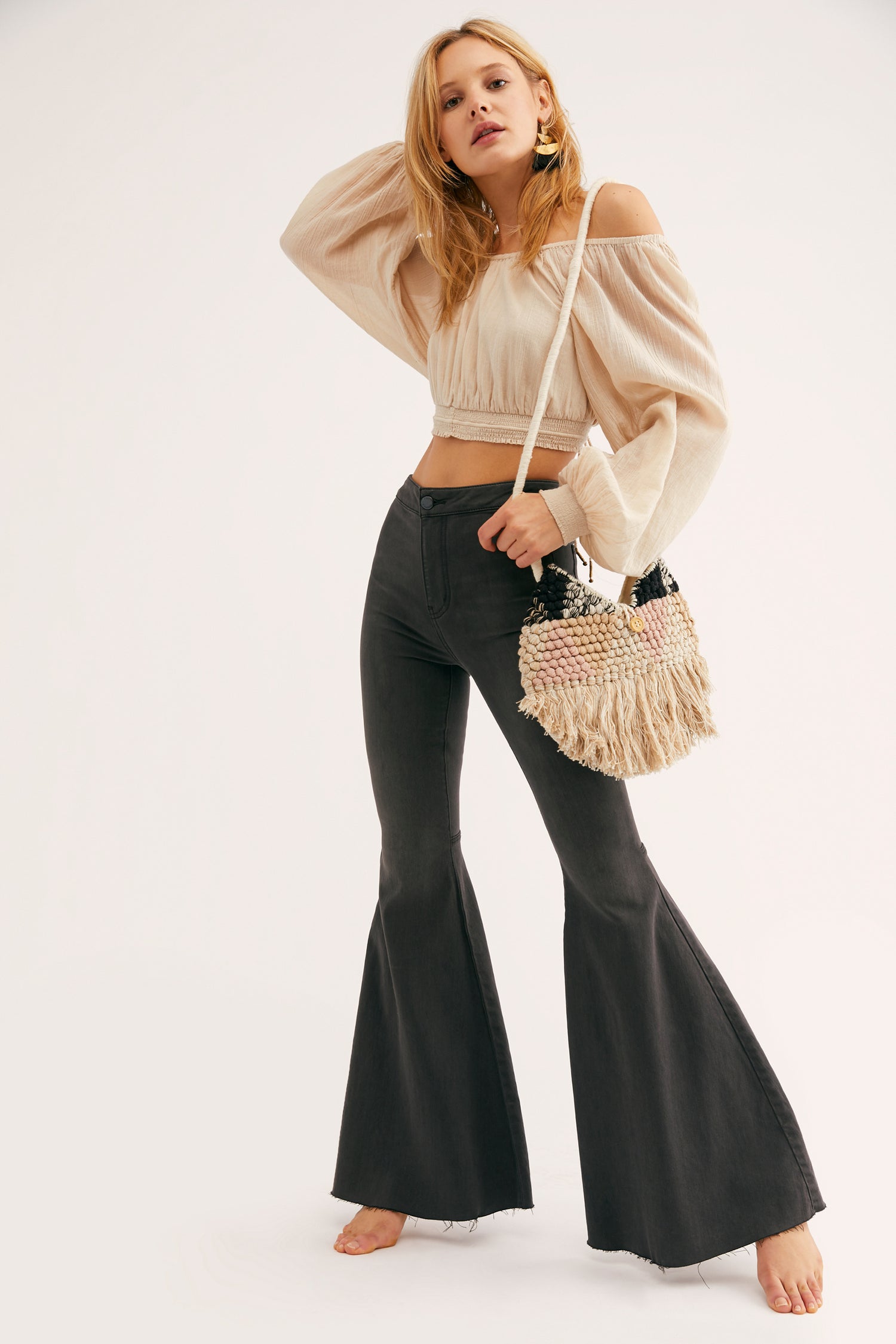 Woman wearing a beige off-shoulder top and black flared pants, holding a woven handbag on a white background.