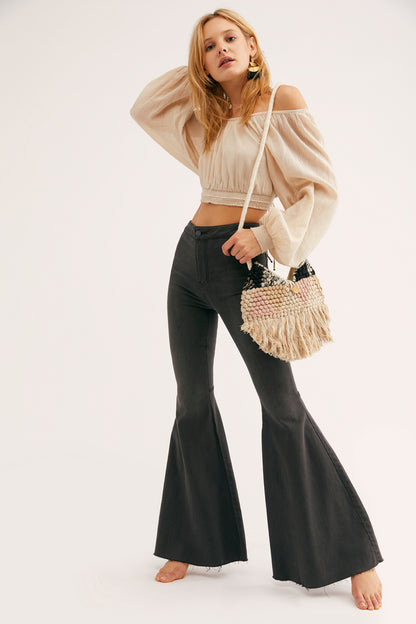 Woman wearing a beige off-shoulder top and black flared pants, holding a woven handbag on a white background.